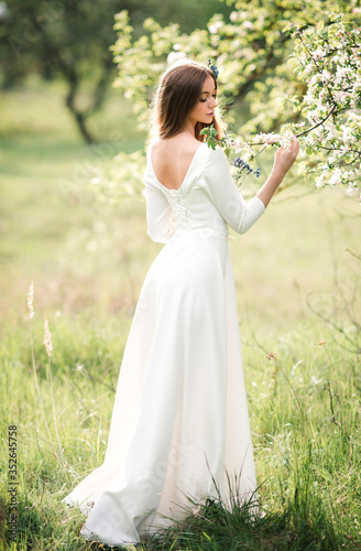 Beautiful young girl with long hair in a white dress with an open back in a summer blooming garden