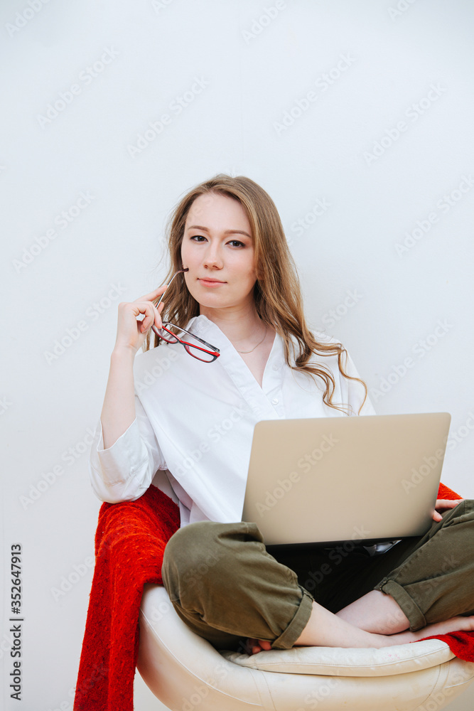 Thoughtful provocative young woman in a white dress-shirt sitting cross-legged on a low spinning chair with laptop, holding glasses next to her face. Thoughtful provocative young woman in a white dress-shirt sitting cross-legged on a low spinning chair with laptop, holding glasses next to her face.