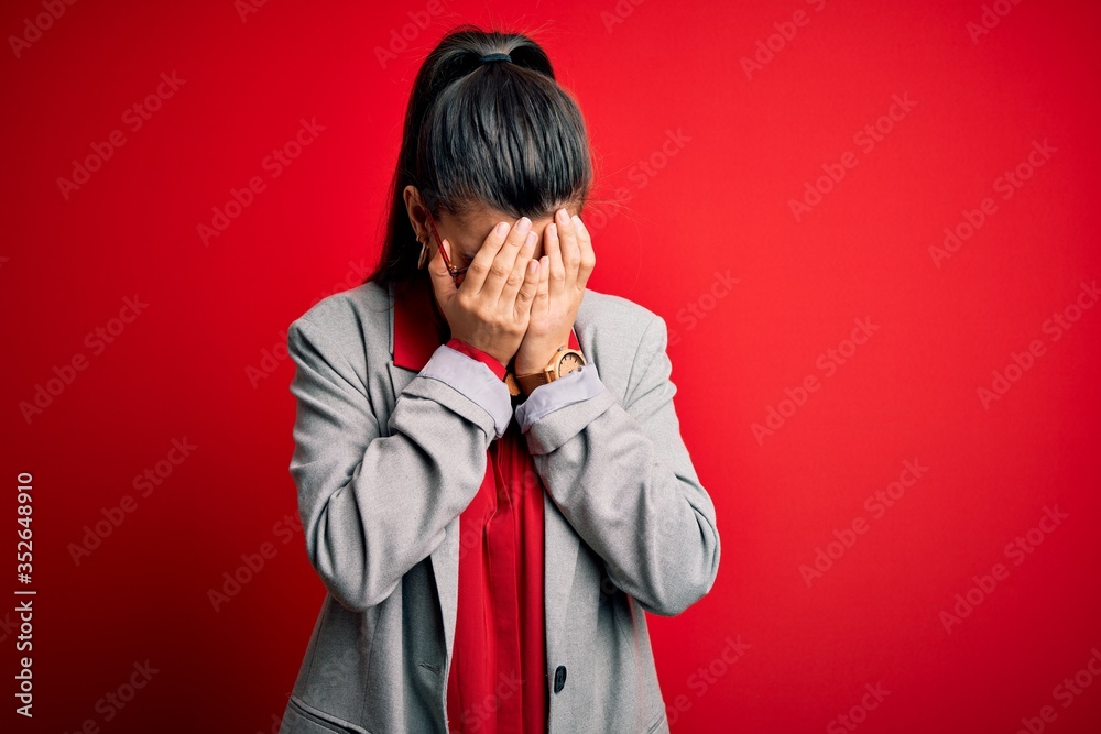 Young beautiful brunette businesswoman wearing jacket and glasses over red background with sad expression covering face with hands while crying. Depression concept. Young beautiful brunette businesswoman wearing jacket and glasses over red background with sad expression covering face with hands while crying. Depression concept.