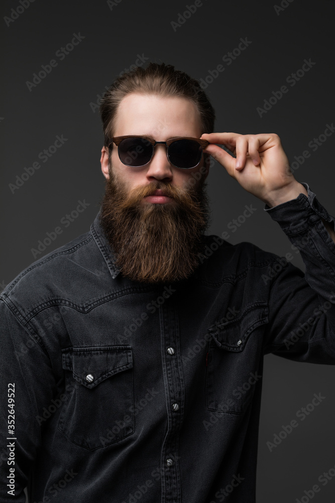 Obraz premium Portrait of handsome s bearded young man with serious expression wearing sunglasses over gray background