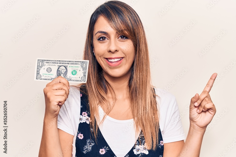 Young beautiful woman holding one dollar smiling happy pointing with hand and finger to the side Young beautiful woman holding one dollar smiling happy pointing with hand and finger to the side
