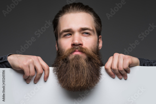 Young long bearded man over blank text on white sheet on gray background.