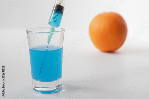 Orange, syringe and glass on a white background. Syringe with blue liquid on the white background. Orange in the background in defocus.