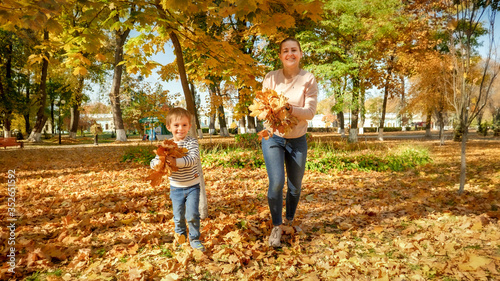 Happy smiling boy with young mother holding autumn leaves and running in park