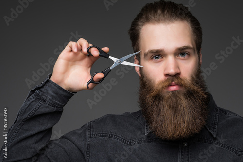 Young bearded man holding barber scissors and razor near his face standing isolated on gray background