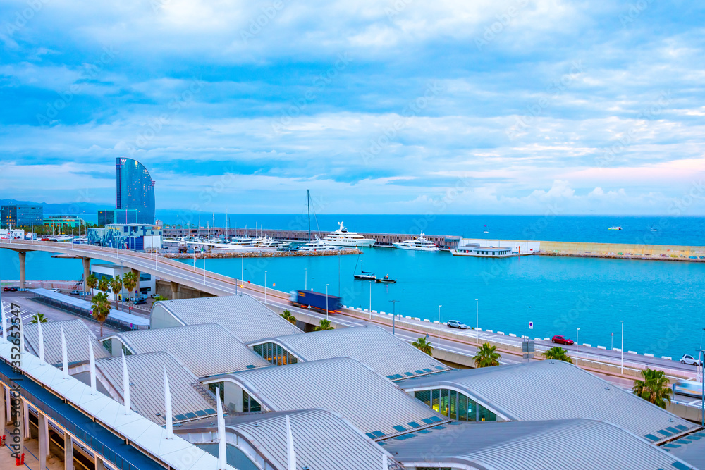 Barcelona, Spain. Morning city skyline view with iconic seaside architecture next to yachts, boats, marine vessels docked in boatyard/ shipyard