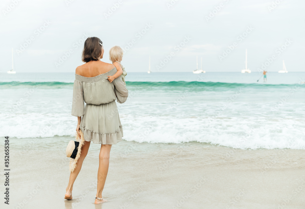 A slender young mother with long legs holds a child with blond hair in her arms and watches the horizon with waves and luxurious white yachts. Vacation at the beach.