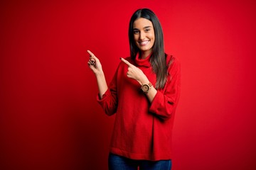 Young beautiful brunette woman wearing casual sweater over isolated red background smiling and looking at the camera pointing with two hands and fingers to the side.