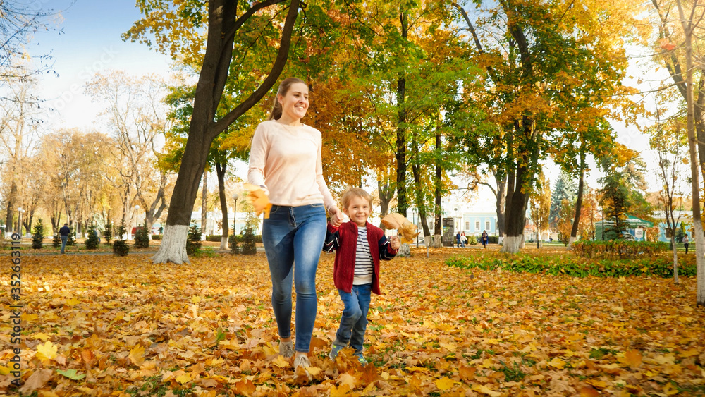Happy laughing little boy walking his mother on yellow fallen leaves at autumn park