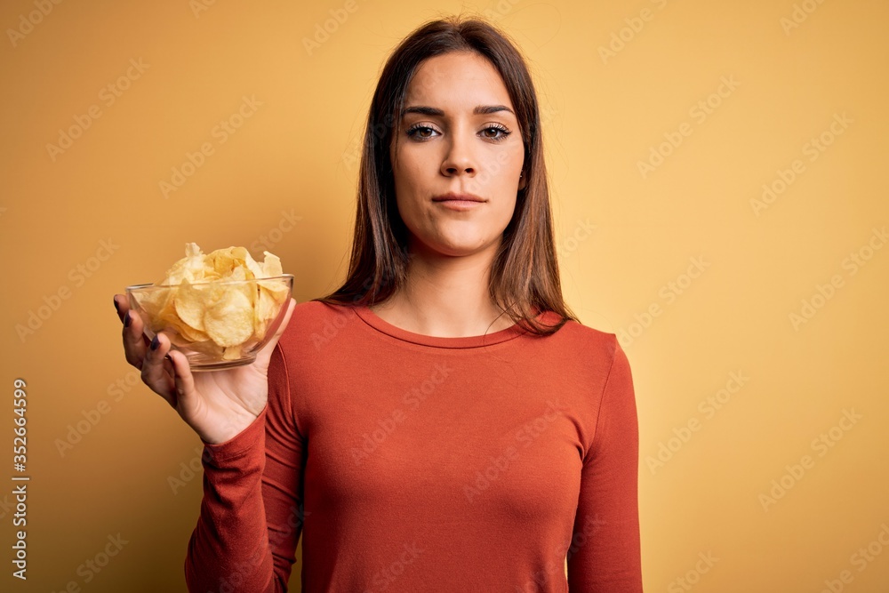 Young beautiful brunette woman holding bowl with chips potatoes over yellow background with a confident expression on smart face thinking serious Young beautiful brunette woman holding bowl with chips potatoes over yellow background with a confident expression on smart face thinking serious
