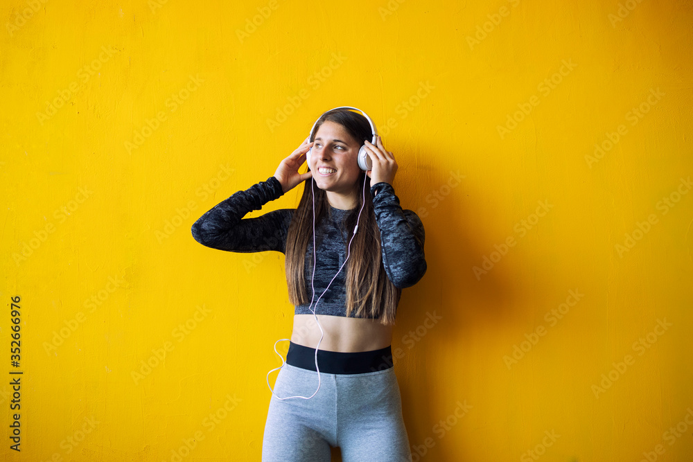 Portrait of sexy muscular build fitness woman standing against yellow background and listening to the music on her headphones. Portrait of sexy muscular build fitness woman standing against yellow background and listening to the music on her headphones.