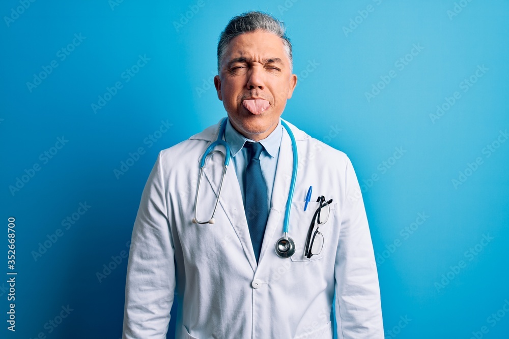 Middle age handsome grey-haired doctor man wearing coat and blue stethoscope sticking tongue out happy with funny expression. Emotion concept. Middle age handsome grey-haired doctor man wearing coat and blue stethoscope sticking tongue out happy with funny expression. Emotion concept.