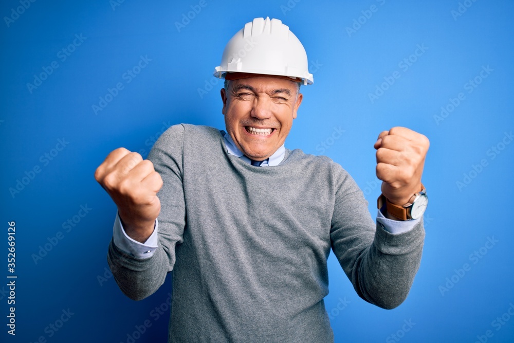 Middle age handsome grey-haired engineer man wearing safety helmet over blue background very happy and excited doing winner gesture with arms raised, smiling and screaming for success. Celebration Middle age handsome grey-haired engineer man wearing safety helmet over blue background very happy and excited doing winner gesture with arms raised, smiling and screaming for success. Celebration