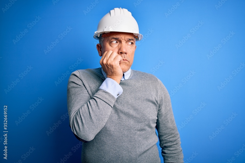 Middle age handsome grey-haired engineer man wearing safety helmet over blue background looking stressed and nervous with hands on mouth biting nails. Anxiety problem. Middle age handsome grey-haired engineer man wearing safety helmet over blue background looking stressed and nervous with hands on mouth biting nails. Anxiety problem.