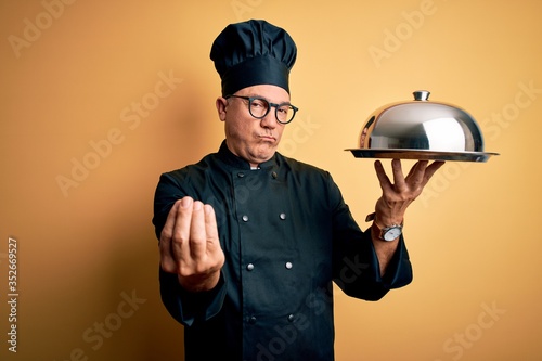 Middle age handsome grey-haired waiter man wearing cooker uniform and hat holding tray doing money gesture with hands, asking for salary payment, millionaire business photo