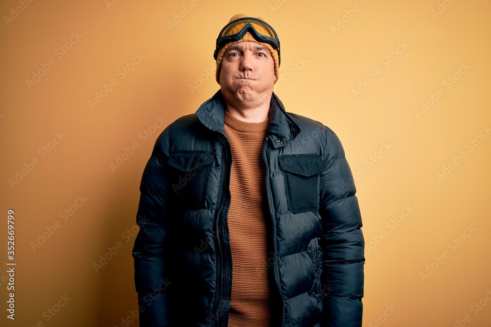 Middle age handsome grey-haired skier man on vacation wearing ski goggles puffing cheeks with funny face. Mouth inflated with air, crazy expression.