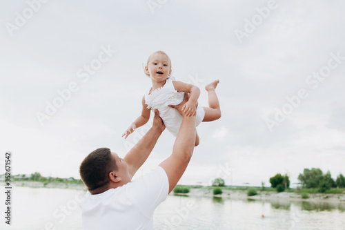 Father throws baby on pier on lake background, girl flies in the sky. Portrait dad with child together. Daddy, little daughter outdoors. Family fishing holiday on pond.