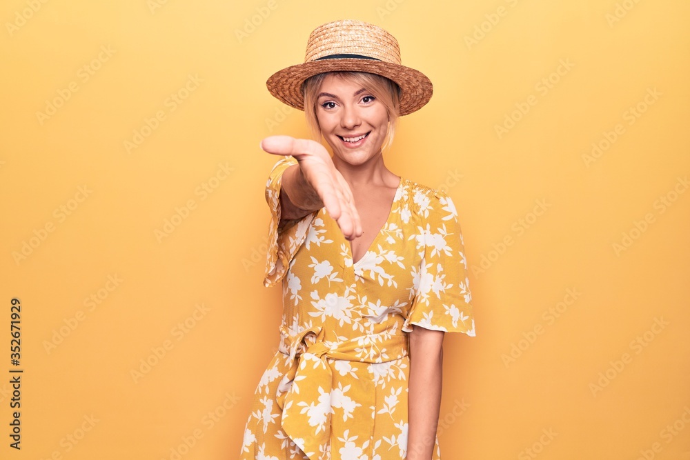 Beautiful blonde woman on vacation wearing summer hat and dress over yellow background smiling friendly offering handshake as greeting and welcoming. Successful business.