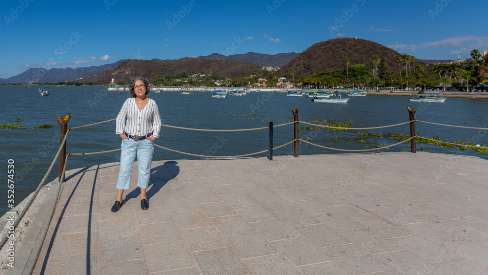 Smiling tourist with casual clothes posing on the boardwalk with the lake with motorboats, the pier and the mountains in the background, sunny day with a clear blue sky in Chapala, Jalisco, Mexico