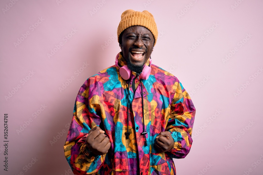 Young handsome african american man wearing colorful coat and cap over pink background very happy and excited doing winner gesture with arms raised, smiling and screaming for success. Celebration