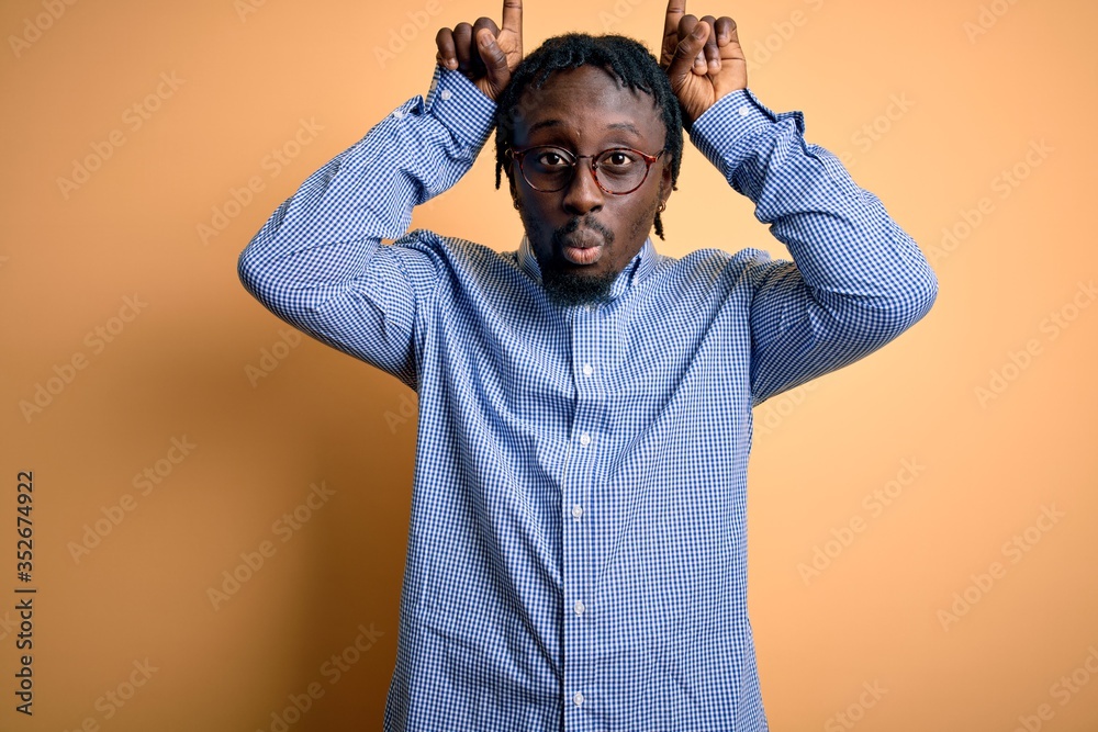 Young handsome african american man wearing shirt and glasses over yellow background doing funny gesture with finger over head as bull horns Young handsome african american man wearing shirt and glasses over yellow background doing funny gesture with finger over head as bull horns