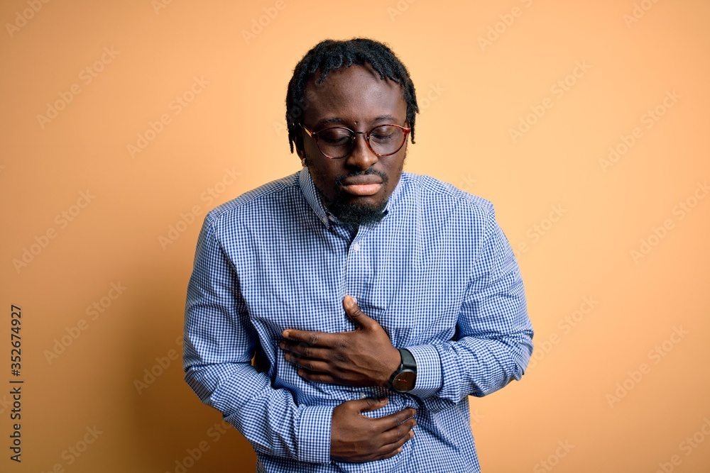 Young handsome african american man wearing shirt and glasses over yellow background with hand on stomach because indigestion, painful illness feeling unwell. Ache concept. Young handsome african american man wearing shirt and glasses over yellow background with hand on stomach because indigestion, painful illness feeling unwell. Ache concept.