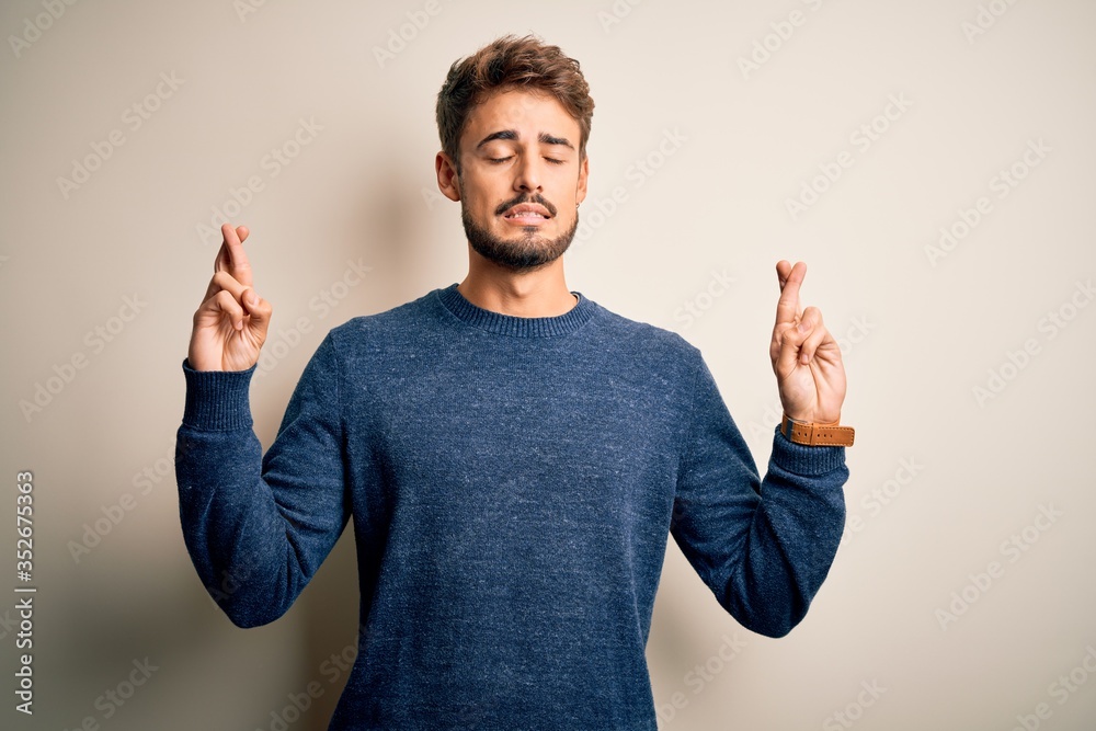 Young handsome man with beard wearing casual sweater standing over white background gesturing finger crossed smiling with hope and eyes closed. Luck and superstitious concept. Young handsome man with beard wearing casual sweater standing over white background gesturing finger crossed smiling with hope and eyes closed. Luck and superstitious concept.