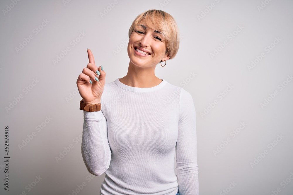 Young beautiful blonde woman with modern short hair hairstyle standing over isolated background showing and pointing up with finger number one while smiling confident and happy. Young beautiful blonde woman with modern short hair hairstyle standing over isolated background showing and pointing up with finger number one while smiling confident and happy.