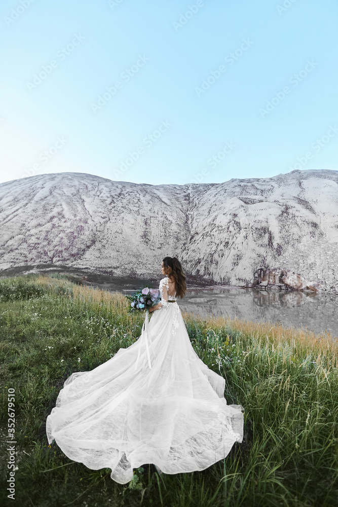 Young woman in bridal dress with a wedding bouquet posing with her back at a beautiful landscape. Wedding fashion concept. Outdoors wedding ceremony.