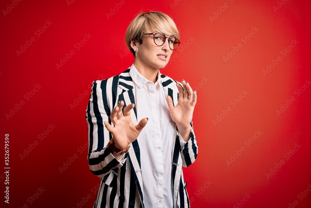 Blonde business woman with short hair wearing glasses and striped jacket over red background disgusted expression, displeased and fearful doing disgust face because aversion reaction