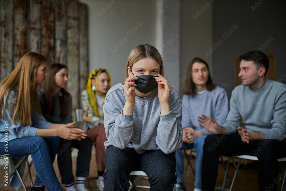 Φωτογραφία young woman afraid of COVID-19, she is wearing medical mask, sits in the center and look at camera while group of people sit in background without masks