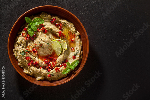 homemade spread of baked eggplant baba ganoush in a bowl with pomegranate seeds, lime, olive oil and lime slices on a black background