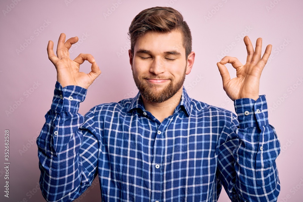 Naklejka premium Young handsome blond man with beard and blue eyes wearing casual shirt standing relax and smiling with eyes closed doing meditation gesture with fingers. Yoga concept.