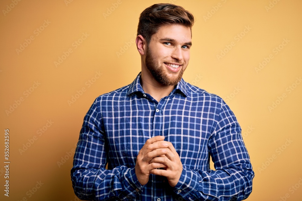 Young blond businessman with beard and blue eyes wearing shirt over yellow background with hands together and crossed fingers smiling relaxed and cheerful. Success and optimistic Young blond businessman with beard and blue eyes wearing shirt over yellow background with hands together and crossed fingers smiling relaxed and cheerful. Success and optimistic