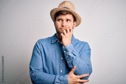 Young handsome blond man with beard and blue eyes wearing denim shirt and summer hat with hand on chin thinking about question, pensive expression. Smiling with thoughtful face. Doubt concept.