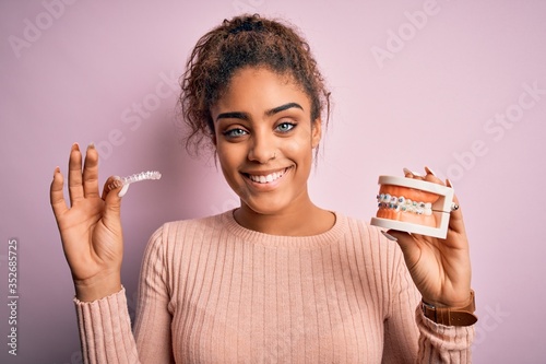 Young african american woman smiling happy holding professional orthodontic denture with metal braces and removable invisible aligner. Comparation of two dental straighten treatments photo
