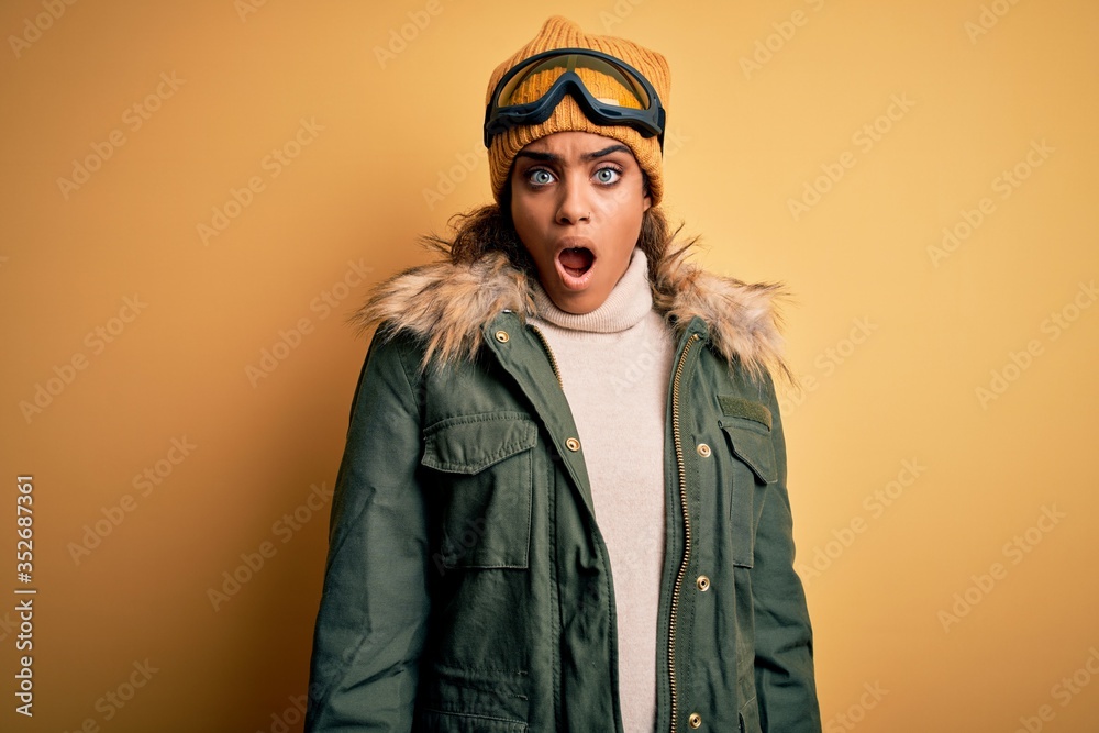 Young african american afro skier girl wearing snow sportswear and ski goggles In shock face, looking skeptical and sarcastic, surprised with open mouth