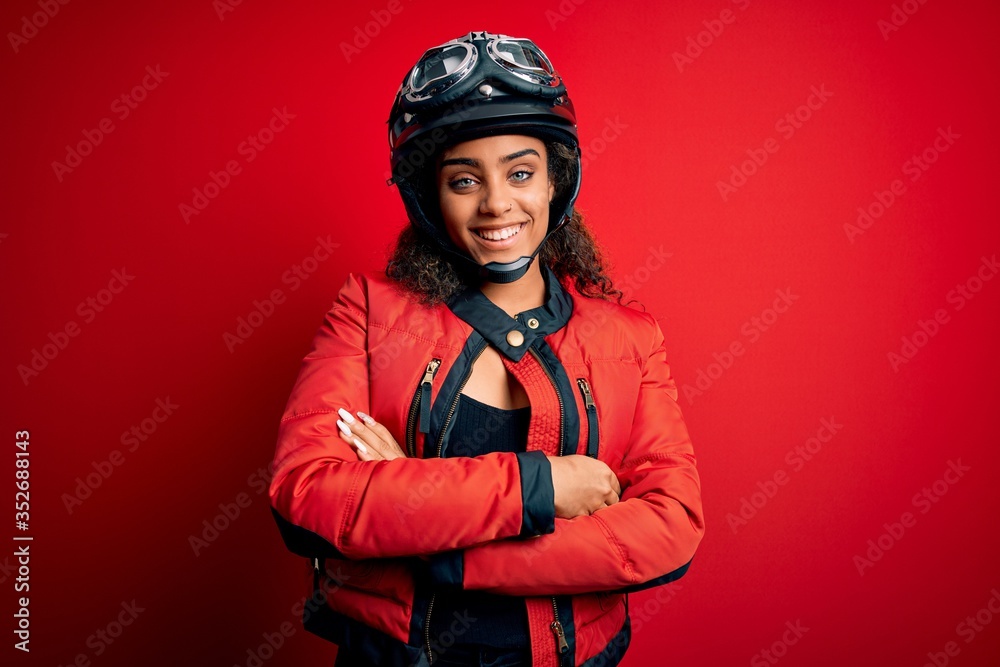 Young african american motorcyclist girl wearing moto helmet and glasses over red background happy face smiling with crossed arms looking at the camera. Positive person. Young african american motorcyclist girl wearing moto helmet and glasses over red background happy face smiling with crossed arms looking at the camera. Positive person.