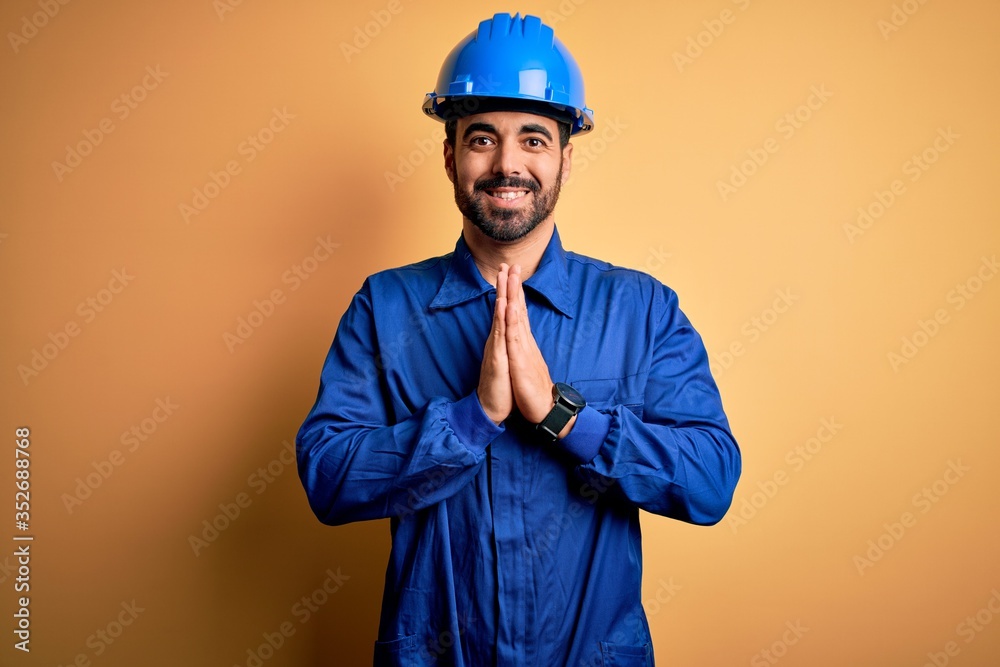 Mechanic man with beard wearing blue uniform and safety helmet over yellow background praying with hands together asking for forgiveness smiling confident.