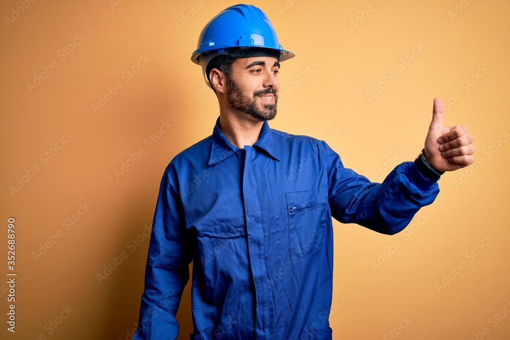 Mechanic man with beard wearing blue uniform and safety helmet over yellow background Looking proud, smiling doing thumbs up gesture to the side