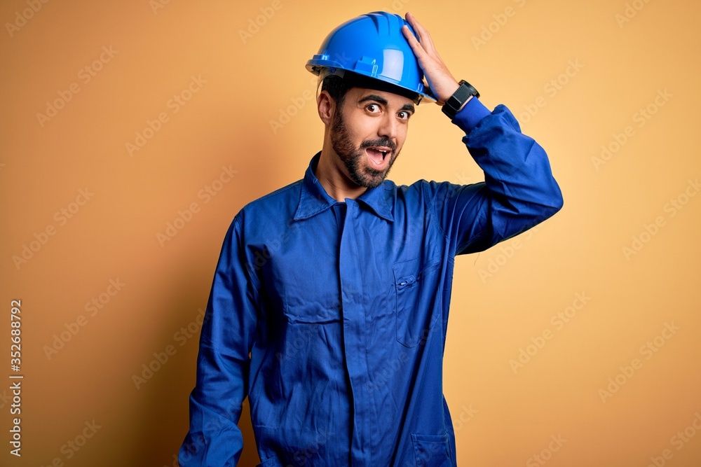 Mechanic man with beard wearing blue uniform and safety helmet over yellow background surprised with hand on head for mistake, remember error. Forgot, bad memory concept. Mechanic man with beard wearing blue uniform and safety helmet over yellow background surprised with hand on head for mistake, remember error. Forgot, bad memory concept.