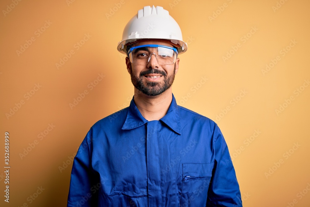 Mechanic man with beard wearing blue uniform and safety glasses over yellow background with a happy and cool smile on face. Lucky person.