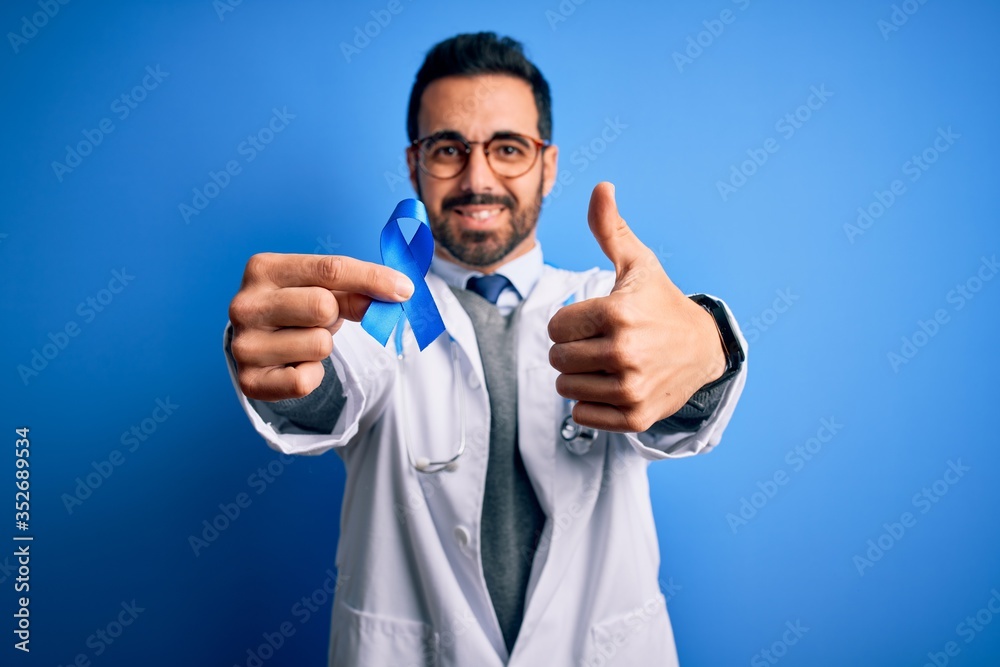 Young handsome doctor man with beard wearing stethoscope holding blue cancer ribbon happy with big smile doing ok sign, thumb up with fingers, excellent sign Young handsome doctor man with beard wearing stethoscope holding blue cancer ribbon happy with big smile doing ok sign, thumb up with fingers, excellent sign