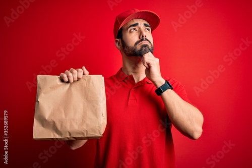 Young handsome delivery man with beard wearing cap holding takeaway paper bag with food serious face thinking about question, very confused idea © Krakenimages.com