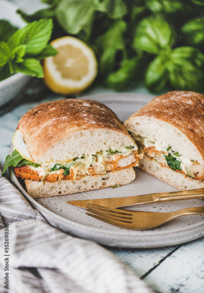 Φωτογραφία Breakfast table with fresh fried fish sandwich with tartare sauce, lemon and arugula cut in halves, fresh greens on plate over rustic wooden table