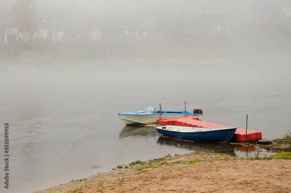 Naklejka premium Sad autumn landscape. Fog and rain over the river. Rescue boats stand at the shore. Walk on the water in bad weather. Two boats moored at the river dock.