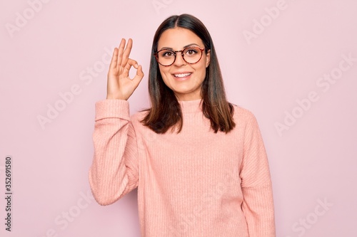 Young beautiful brunette woman wearing casual sweater and glasses over pink background smiling positive doing ok sign with hand and fingers. Successful expression.