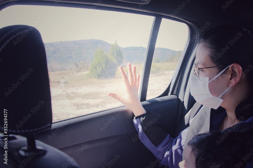 Mujer joven con máscara médica de protección viaja de pasajero en un automóvil junto a su bebé durante la pandemia. Mujer con tapabocas observa desde el interior de un automóvil con la mano en la vent