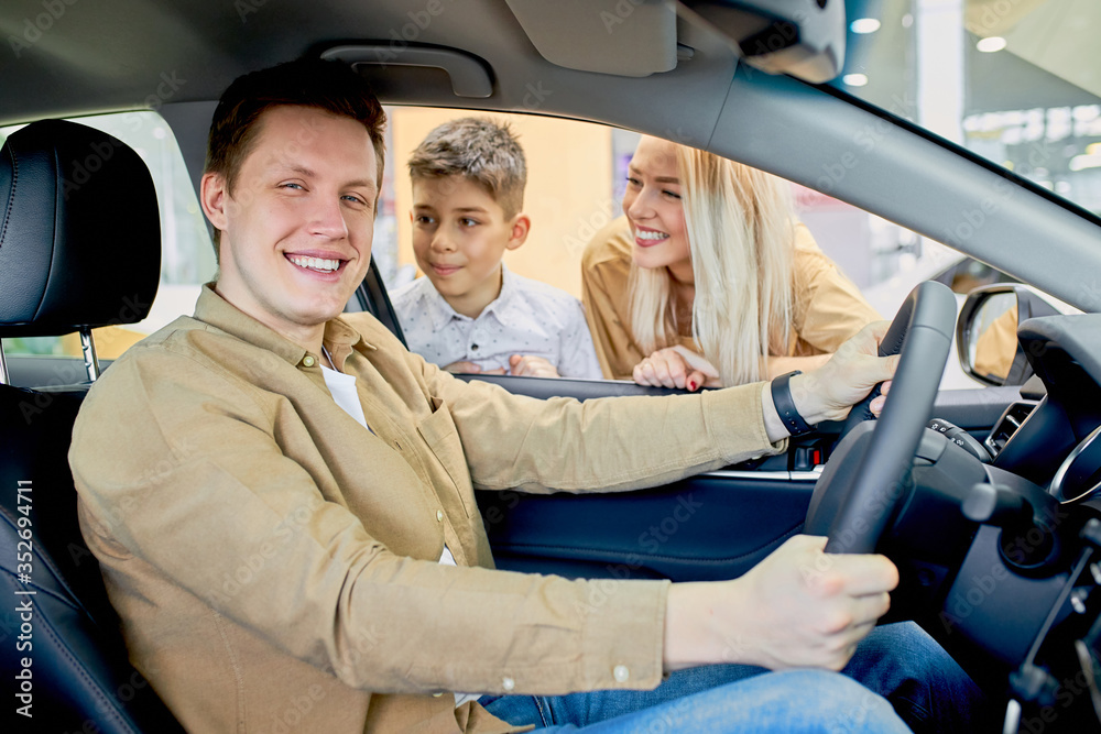 smiling young caucasian car owner in dealership, positive guy sit at front seat of the car, likes design of auto, wife and son waiting for his reaction on car, ask about feelings