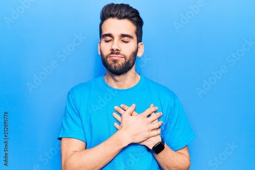Young handsome man with beard wearing casual t-shirt smiling with hands on chest, eyes closed with grateful gesture on face. health concept.